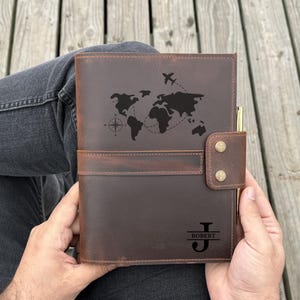 May include: A brown leather journal with a world map design and an airplane graphic. The journal has a pen holder and a snap closure. The initials "J" and "ROBERT" are engraved on the cover.