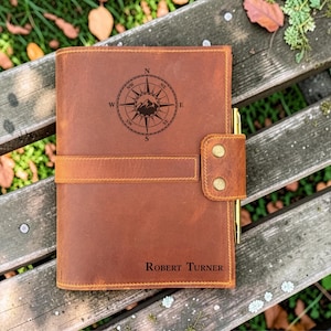 May include: A tan leather journal featuring a dark brown engraved compass design with mountain peaks in the centre. The cover includes a horizontal strap secured by two brass-coloured metal snaps and a gold-toned pen held in a side loop. The name Robert Turner is engraved in the bottom right corner.