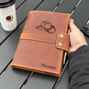 May include: A tan leather journal cover featuring a dark brown engraved cowboy hat graphic on the front cover and the name Thomas engraved in the bottom right corner. The cover includes a horizontal strap with two brass-coloured metal snap buttons.