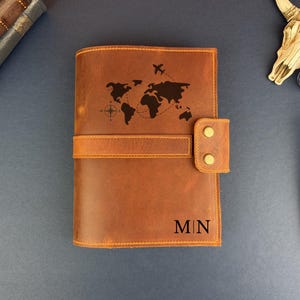 May include: A brown leather journal featuring a laser-engraved world map, a compass rose, and a small airplane icon. The cover includes a horizontal strap with two brass-colored snap buttons and the initials M|N engraved in the bottom right corner.