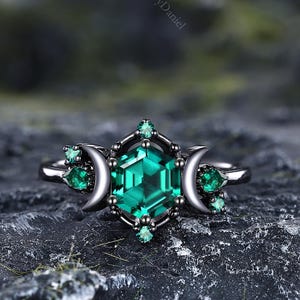 May include: A silver ring with a large, emerald-cut green gemstone at the centre, encircled by smaller green gems and crescent moon details. The ring has a dark, oxidised finish, set against a textured, dark grey background.