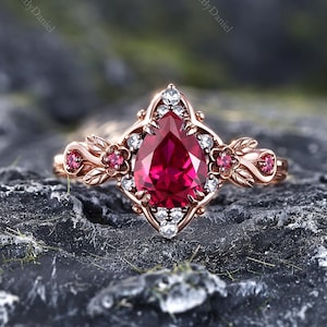 May include: A rose gold ring featuring a central pear-shaped red gemstone surrounded by small white accent stones. The band is decorated with leaf-like metal motifs and small round red stones on each side of the central setting.