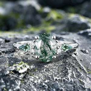 May include: A silver ring featuring a large, oval-cut moss agate gemstone with green inclusions. The ring is accented with small, round diamonds and triangular green gemstones on either side of the main stone. The ring is set on a textured, gray surface.