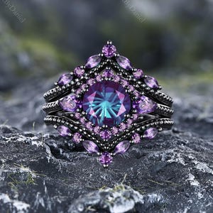 May include: A black metal ring featuring a large, round, color-changing center stone in shades of blue and purple. The ring is surrounded by a geometric frame of smaller, round and marquise-cut purple stones, creating a layered, ornate appearance.