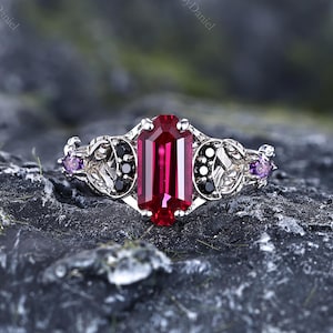 May include: A silver-toned ring featuring a central emerald-cut deep red gemstone. The band is adorned with intricate metalwork, small black circular stones, and two small purple accent stones on the outer edges of the decorative setting.