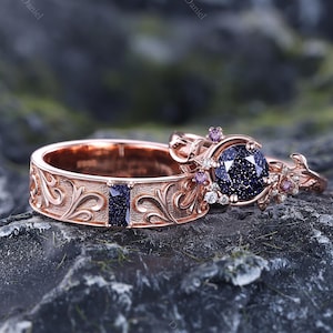 May include: Two rose gold rings. One ring features a large, round, dark blue gemstone with smaller purple and white stones. The other ring has a rectangular dark blue stone and ornate floral detailing. The rings are set against a dark, textured background.
