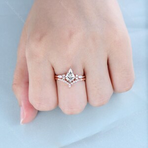 May include: A rose gold engagement ring set with a square-shaped diamond center stone and smaller diamonds on the band. The ring is on a finger.