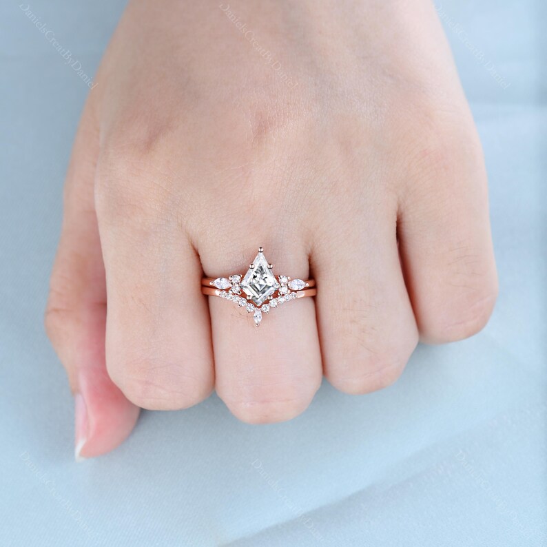 May include: A rose gold engagement ring set with a square-shaped diamond center stone and smaller diamonds on the band. The ring is on a finger.