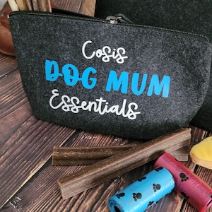 May include: A dark gray felt pouch with the text "Dog Mum Essentials" in white and blue. The pouch is open, revealing dog treats. Rolls of blue and red dog waste bags and more treats are scattered around the pouch on a wooden surface.
