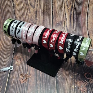 Op de afbeelding: Een set van tien kleurrijke polsbandjes met witte tekst. De polsbandjes zijn gemaakt van satijnen lint en hebben een zwarte rand. De tekst op de polsbandjes omvat "Prost Mad", "Trauzeugen", "Cheers! To", "Braut", "Girls Trip", "Team Bride", "Verena", "The Bride", "Agesellen" en "The Bride".