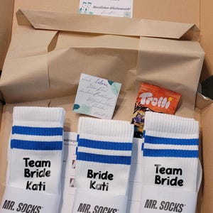Könnte beinhalten: Ein offener Karton mit drei Paar weißen Socken mit blauen Streifen und den Aufschriften "Team Bride Kati" und "Bride Kati". Die Socken sind von "MR. SOCKS". Eine Karte und Süßigkeiten sind ebenfalls enthalten.