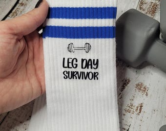 May include: White athletic socks with blue stripes and the text "LEG DAY SURVIVOR" and a dumbbell graphic. These socks are designed for fitness enthusiasts and gym-goers.