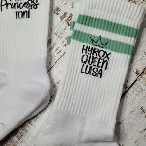 Könnte beinhalten: Zwei weiße Sportsocken mit gestreiften Bündchen. Eine Socke hat den Aufdruck "PILATES Princess TONI" in Schwarz, die andere "HYROX QUEEN LUISA" mit einer Kronengrafik. Die Bündchen sind lila und grün.