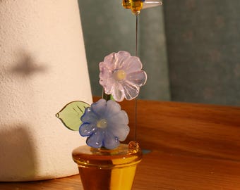 May include: A glass flower arrangement in a yellow pot with a blue dragonfly on top. The flowers are pink, blue, and green.
