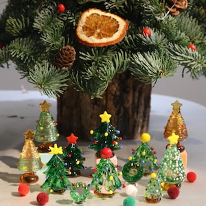 May include: A collection of 10 small glass Christmas tree decorations in various colours and designs, including green, red, white, and gold. The decorations are arranged on a white surface with red and green pom-poms.