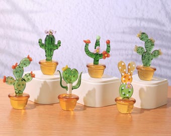 May include: Six glass cactus figurines in various styles, including a traditional cactus, a prickly pear cactus, and a bunny-shaped cactus. The figurines are all green and have small, colorful flowers. They are displayed on a white surface.