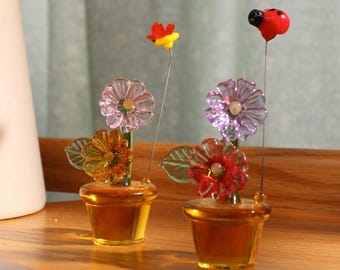 May include: Two glass flower arrangements in pots. The flowers are pink, orange, and red. One arrangement has a red ladybug on top and the other has a yellow butterfly.