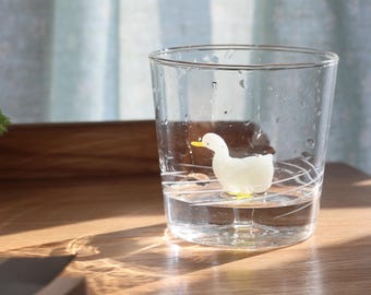Könnte beinhalten: Ein klares Glas mit einer weißen Entenfigur im Inneren, gefüllt mit Wasser. Die Ente hat einen gelben Schnabel und gelbe Füße. Das Glas hat ein schlichtes, elegantes Design mit ein paar weißen Linien. Das Glas steht auf einer Holzoberfläche.