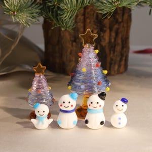 May include: Five glass Christmas decorations, including two blue and purple Christmas trees and three white snowmen with blue hats and scarves. The snowmen have black eyes and smiling mouths.