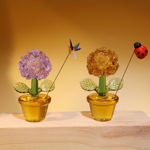 May include: Two glass flower sculptures in pots. The flower on the left is purple and has a blue and white dragonfly perched on it. The flower on the right is yellow and has a red ladybug perched on it.
