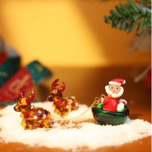 May include: A glass Christmas ornament set featuring Santa Claus in a green sleigh pulled by two reindeer. The reindeer are brown with white spots and have red antlers. The sleigh is decorated with a red bow and a gold chain.