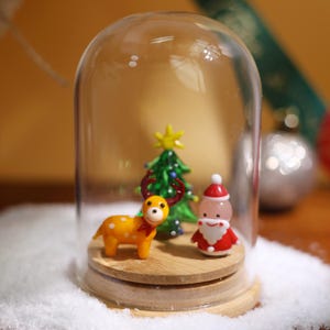 May include: A glass dome with a wooden base contains a miniature Christmas scene. A yellow and white reindeer figurine stands next to a red and white Santa Claus figurine. A green glass Christmas tree with a yellow star sits in the center.