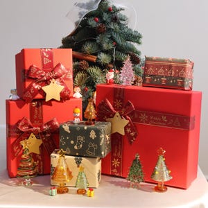 Könnte beinhalten: Rote und grüne Geschenkboxen mit goldenen Bändern und Sternendekorationen. Die Boxen sind mit Weihnachtsmotiven verziert. Auf dem Tisch stehen kleine gläserne Weihnachtsbäume.