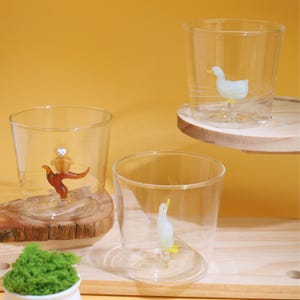 May include: Three clear glass tumblers with different glass animal figurines inside. The tumblers are sitting on wooden coasters. The animals are a brown owl perched on a branch, a white duck, and a white penguin.