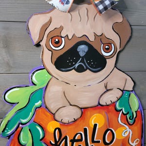 May include: A wooden pug dog decoration sitting on a pumpkin with the words "hello Pumpkin". The pumpkin is orange with green leaves and purple accents. A bow with pumpkins and a black and white checkered pattern is at the top.
