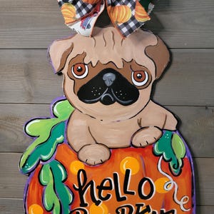 May include: A wooden pug dog decoration sitting on a pumpkin with the words "hello pumpkin" in black script. The pumpkin is orange with yellow spots and green leaves. A decorative bow with paw prints and pumpkins is at the top.