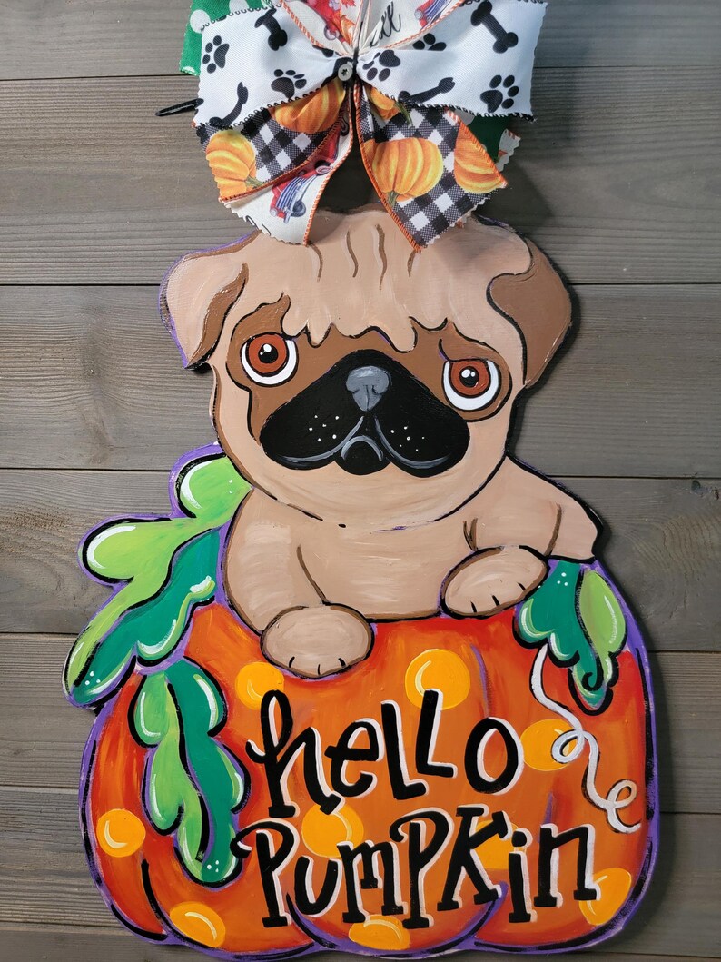 May include: A wooden pug dog decoration sitting on a pumpkin with the words "hello pumpkin" in black script. The pumpkin is orange with yellow spots and green leaves. A decorative bow with paw prints and pumpkins is at the top.