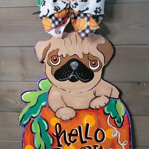 May include: A wooden pug dog decoration sitting on a pumpkin with the words "hello pumpkin". The pumpkin is orange with green leaves and purple outlines. A decorative bow with pumpkins, paw prints, and red trucks tops the decoration.