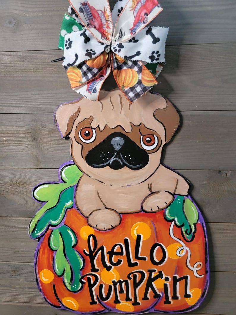 May include: A wooden pug dog decoration sitting on a pumpkin with the words "hello pumpkin". The pumpkin is orange with green leaves and purple outlines. A decorative bow with pumpkins, paw prints, and red trucks tops the decoration.