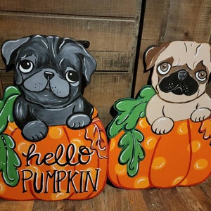 May include: Two wooden pug dog decorations. One is black, the other is tan. Both are sitting in orange pumpkins with green leaves. The orange pumpkins have the words "hello PUMPKIN" painted on them in white script.