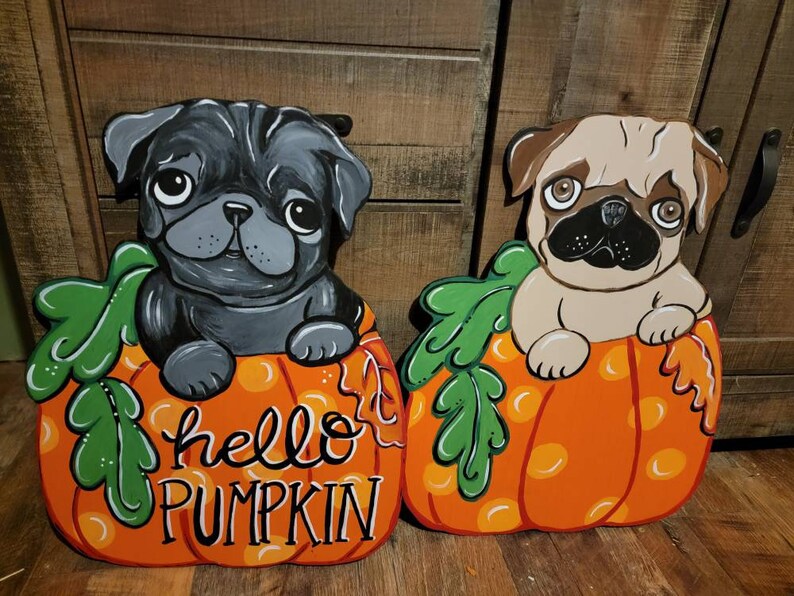 May include: Two wooden pug dog decorations. One is black, the other is tan. Both are sitting in orange pumpkins with green leaves. The orange pumpkins have the words "hello PUMPKIN" painted on them in white script.