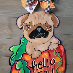 May include: A wooden fall-themed door hanger featuring a pug dog sitting on a pumpkin. The pumpkin is orange and has the words "hello Fall" written on it. A decorative bow with pumpkins and paw prints tops the hanger.