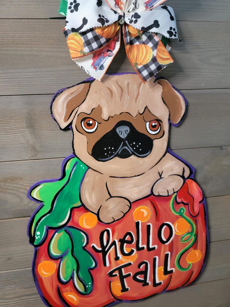 May include: A wooden fall-themed door hanger featuring a pug dog sitting on a pumpkin. The pumpkin is orange and has the words "hello Fall" written on it. A decorative bow with pumpkins and paw prints tops the hanger.