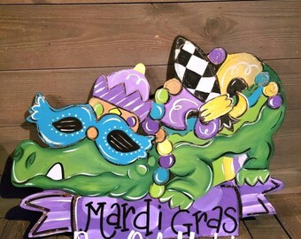 May include: A green alligator wearing a purple, blue, and gold Mardi Gras mask and hat. The alligator is sitting on a purple banner with the text "Mardi Gras Barn Owl Market".