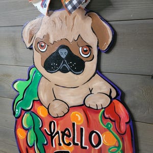 May include: A wooden fall-themed decoration featuring a pug dog sitting on a pumpkin. The pumpkin is orange with green vines and the words "hello Fall". A decorative bow with pumpkin and plaid patterns is at the top.