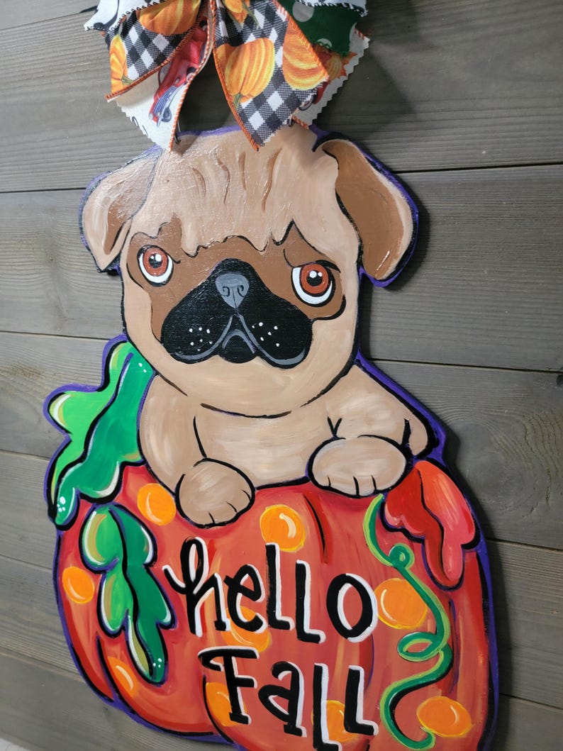 May include: A wooden fall-themed decoration featuring a pug dog sitting on a pumpkin. The pumpkin is orange with green vines and the words "hello Fall". A decorative bow with pumpkin and plaid patterns is at the top.