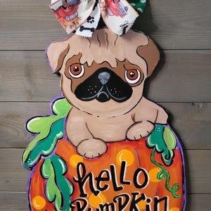 May include: A wooden pug dog decoration sitting in a pumpkin with the words "hello pumpkin" painted on it. The pumpkin is orange with green leaves and a purple outline. A decorative bow with dog bones and cars is attached to the top.