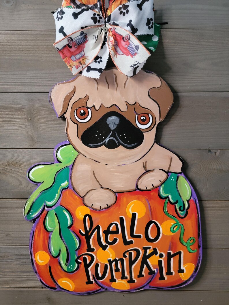 May include: A wooden pug dog decoration sitting in a pumpkin with the words "hello pumpkin" painted on it. The pumpkin is orange with green leaves and a purple outline. A decorative bow with dog bones and cars is attached to the top.