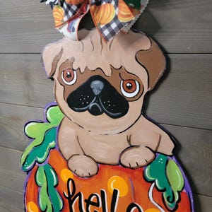 May include: A wooden pug dog decoration sitting on top of a pumpkin with the words "hello PUMPKIN" painted on it. The pumpkin is orange with green leaves and a purple outline. A decorative bow with paw prints and pumpkins is attached to the top.
