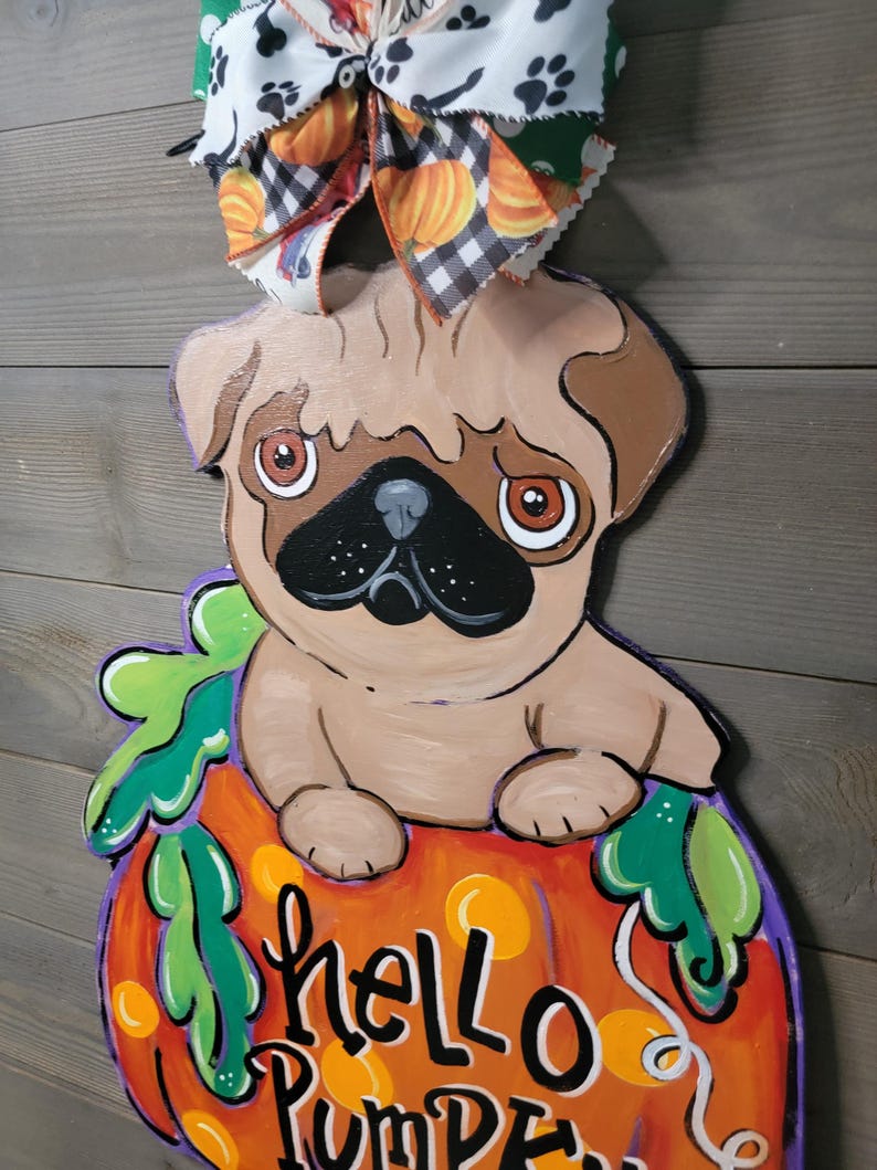 May include: A wooden pug dog decoration sitting on top of a pumpkin with the words "hello PUMPKIN" painted on it. The pumpkin is orange with green leaves and a purple outline. A decorative bow with paw prints and pumpkins is attached to the top.