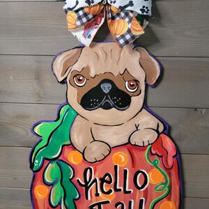 May include: A wooden fall-themed door hanger featuring a pug dog sitting on a pumpkin. The pumpkin is painted red with the words "hello Fall" in white. A decorative bow with pumpkins, paw prints, and trucks tops the design.
