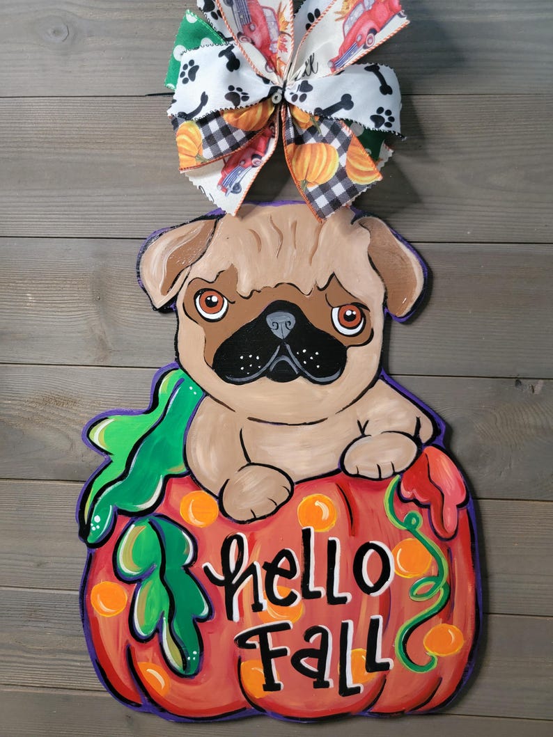 May include: A wooden fall-themed door hanger featuring a pug dog sitting on a pumpkin. The pumpkin is painted red with the words "hello Fall" in white. A decorative bow with pumpkins, paw prints, and trucks tops the design.