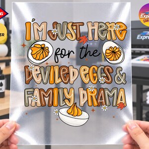 May include: A clear DTF transfer sheet featuring the text I'M JUST HERE for the DEVILED EGGS & FAMILY DRAMA in a retro, multi-colored font. The design includes illustrations of halved deviled eggs, small white daisies, and orange autumn leaves.