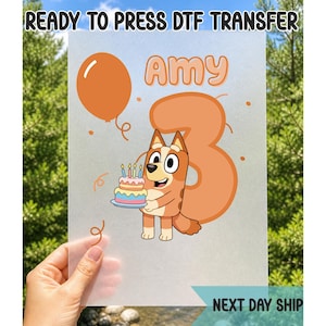 May include: A clear DTF transfer sheet featuring a cartoon dog holding a birthday cake with candles. The design includes the name "Amy" and the number "3" in orange. The text "READY TO PRESS DTF TRANSFER" and "NEXT DAY SHIP" are also visible.