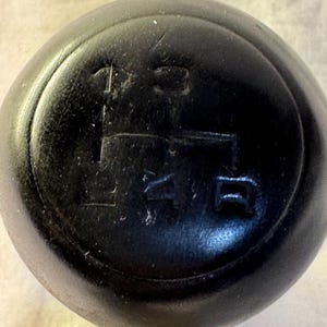 May include: A black gear stick knob displaying a 5-speed transmission layout. The numbers 1, 2, 3, 4, and R are embossed, showing the gear positions. The knob is circular and seems to be made from a hard-wearing material.