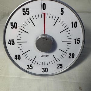May include: A round, white kitchen timer with a silver rim. The timer has black numbers and markings, with a red hand indicating the time. The timer has a silver knob in the center and the brand name "Lavigo" is printed on the face.